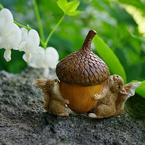 Two squirrel figurines holding a large acorn on a rock with flowers in the background.